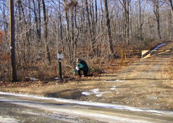 Looking west from the trail. Shown are the powerpole, sign and gated woods road. Rich points to the mark.