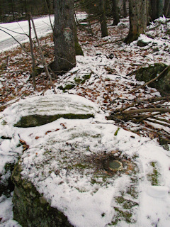 The mark is set in top and in approximately the center of the boulder. A blazed tree can be seen just beyond the mark.