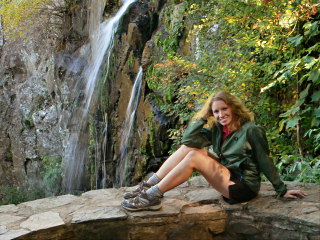 Zhanna sits on a cold stone wall along the trail, with Lewis Falls in the background.